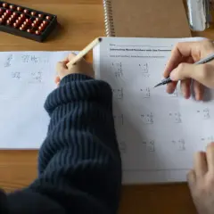 Dziecko rozwiązuje zadania matematyczne, ćwicząc odejmowanie ułamków. Obok leży liczydło, a na stole zeszyt z zapisanym ciągiem arytmetycznym.