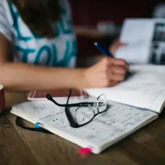 Kobieta pisze w zeszycie, obok kubek kawy i okulary. To idealne warunki, by nauczyć się języka obcego samemu.