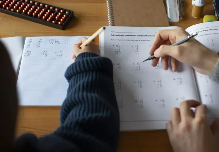 Dziecko rozwiązuje zadania matematyczne, ćwicząc odejmowanie ułamków. Obok leży liczydło, a na stole zeszyt z zapisanym ciągiem arytmetycznym.