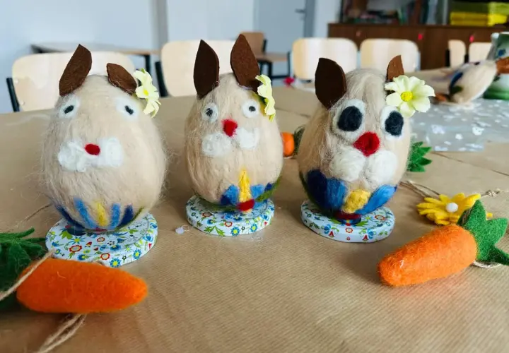 Trzy filcowe zajączki, ozdobione kwiatkami i kolorowymi spódniczkami, jakby mówiły po kaszubsku. Obok leżą filcowe marchewki.
