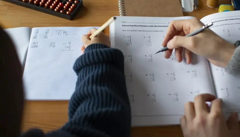 Dziecko rozwiązuje zadania matematyczne, ćwicząc odejmowanie ułamków. Obok leży liczydło, a na stole zeszyt z zapisanym ciągiem arytmetycznym.