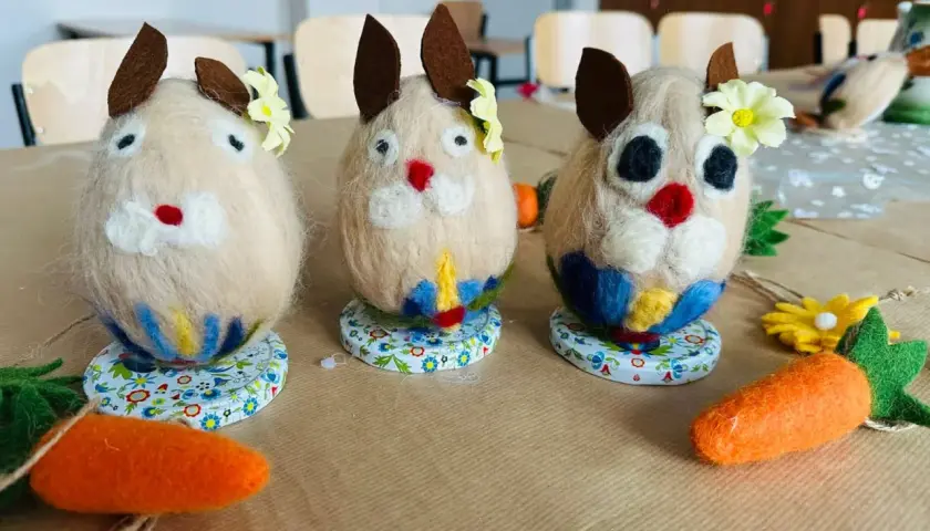 Trzy filcowe zajączki, ozdobione kwiatkami i kolorowymi spódniczkami, jakby mówiły po kaszubsku. Obok leżą filcowe marchewki.