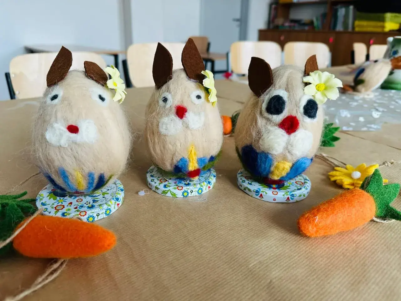 Trzy filcowe zajączki, ozdobione kwiatkami i kolorowymi spódniczkami, jakby mówiły po kaszubsku. Obok leżą filcowe marchewki.