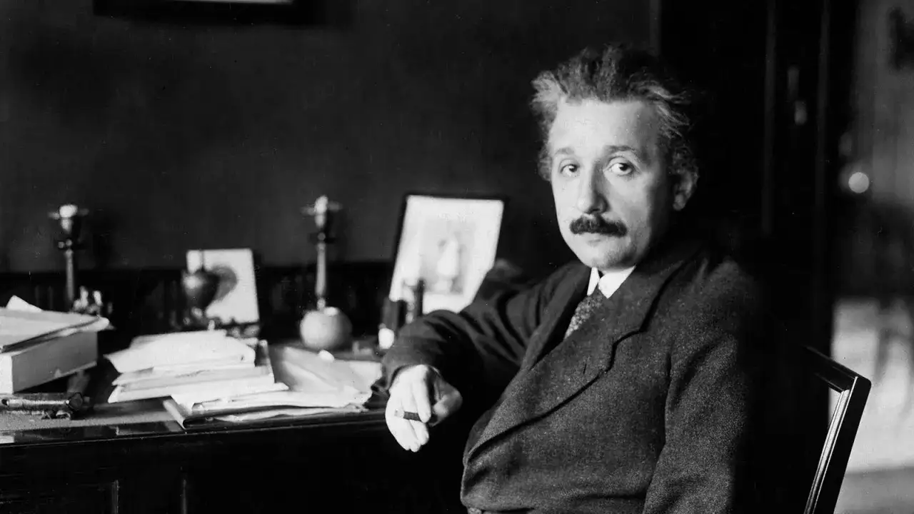 Albert Einstein, znany z tego, że co wynalazł teorię względności, siedzi przy biurku pełnym papierów.