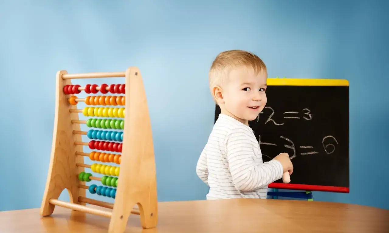 Uśmiechnięty chłopiec przy tablicy z prostymi równaniami, obok kolorowe liczydło. To świetna matematyka w praktyce dla najmłodszych.