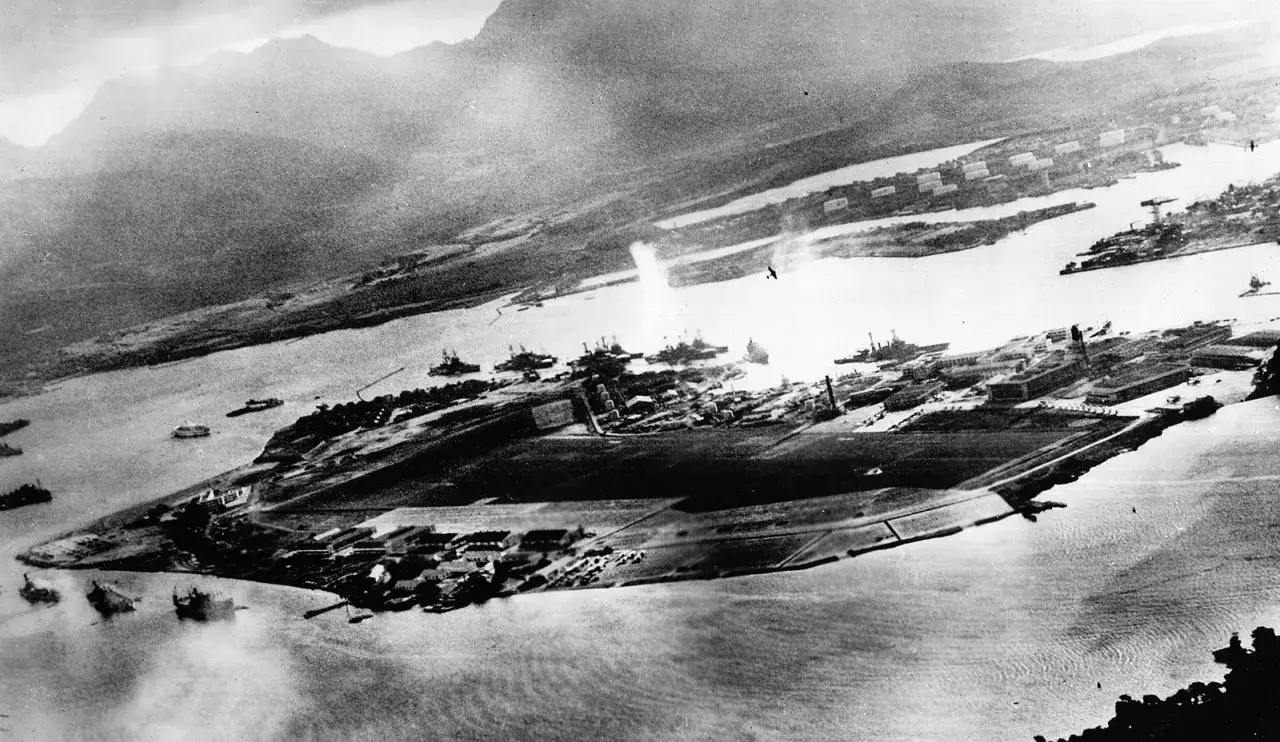 Widok z lotu ptaka na Pearl Harbor podczas ataku. Dym unosi się z okrętów, ukazując skutki japońskiego ataku, który był jedną z przyczyn przystąpienia USA do II wojny światowej.